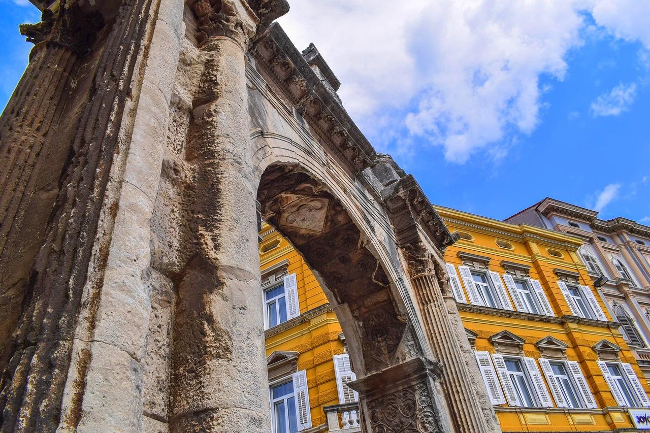 Starożytny kamienny łuk, detale architektoniczne, żółty budynek, błękitne niebo.
