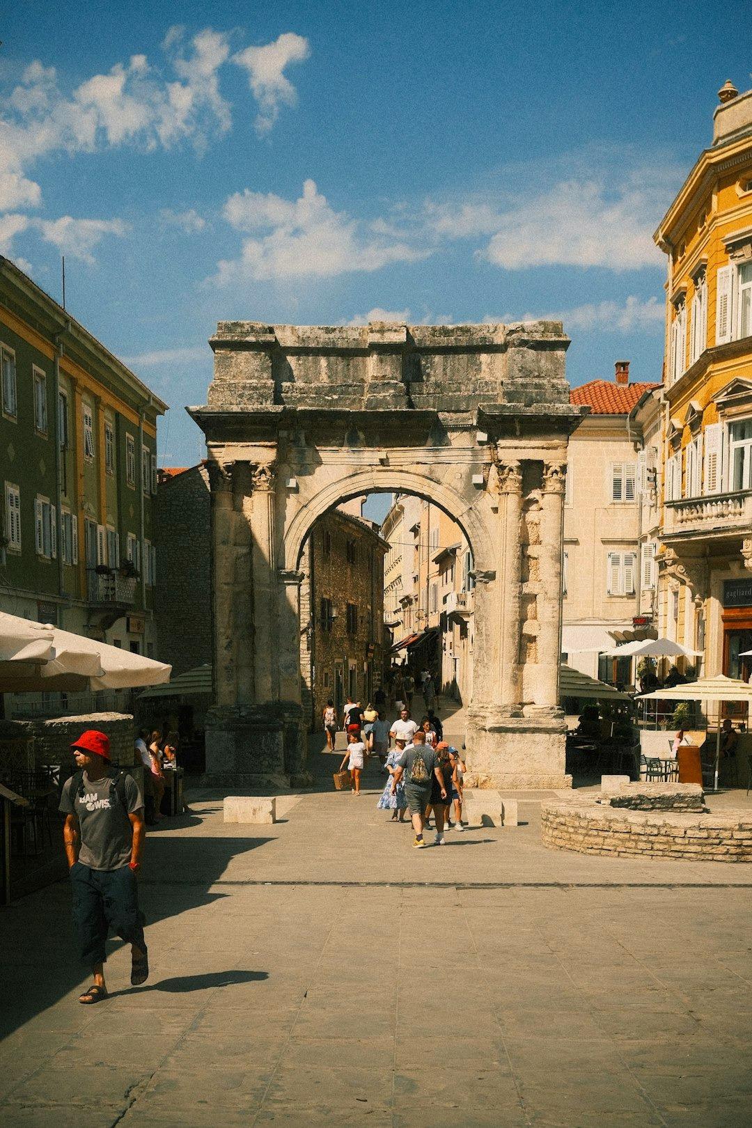 Zabytkowy łuk i przechodnie na słonecznym placu miasta.