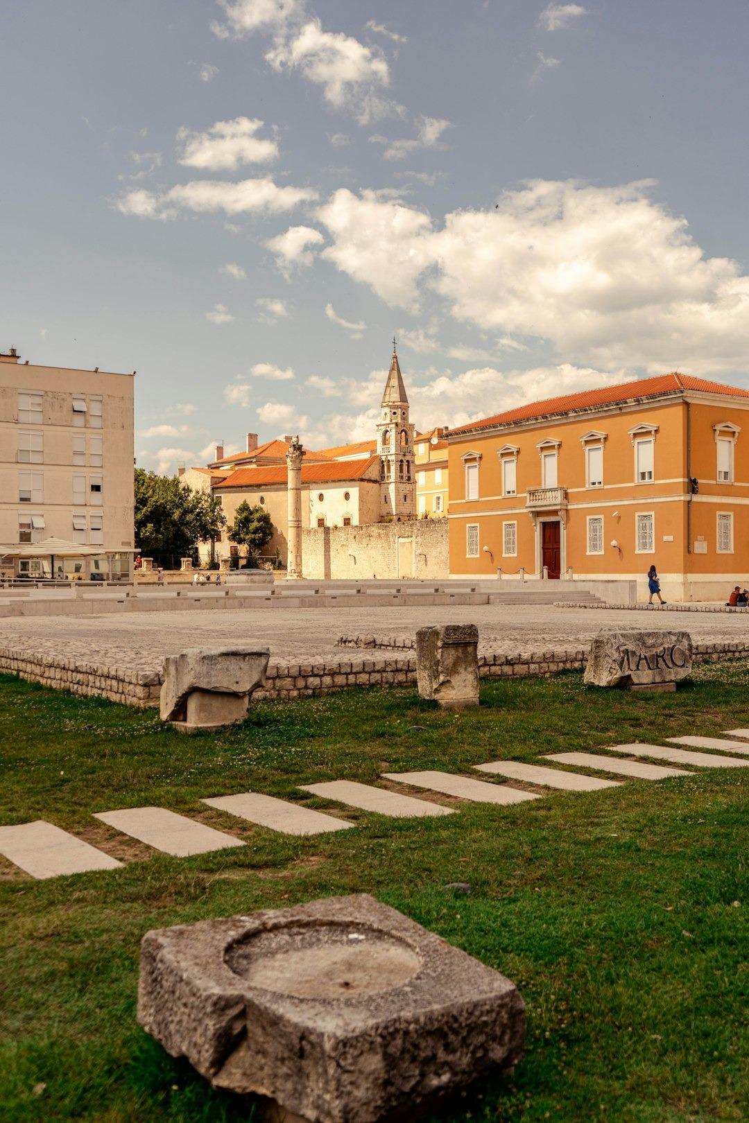 Historyczny plac ze starożytnymi ruinami, dzwonnicą i pomarańczowym budynkiem w słoneczny dzień.