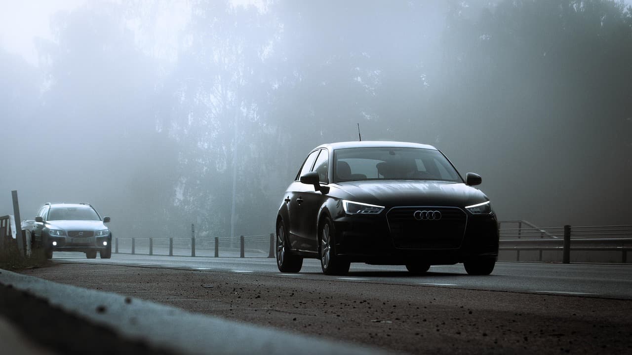 Czarne Audi i Volvo jadą mglistą drogą, włączone światła.