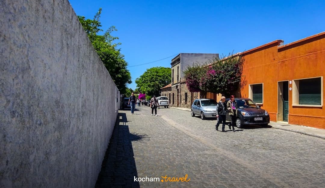 Słoneczna brukowana ulica, stare miasto, kolorowe budynki, spacerujący ludzie.