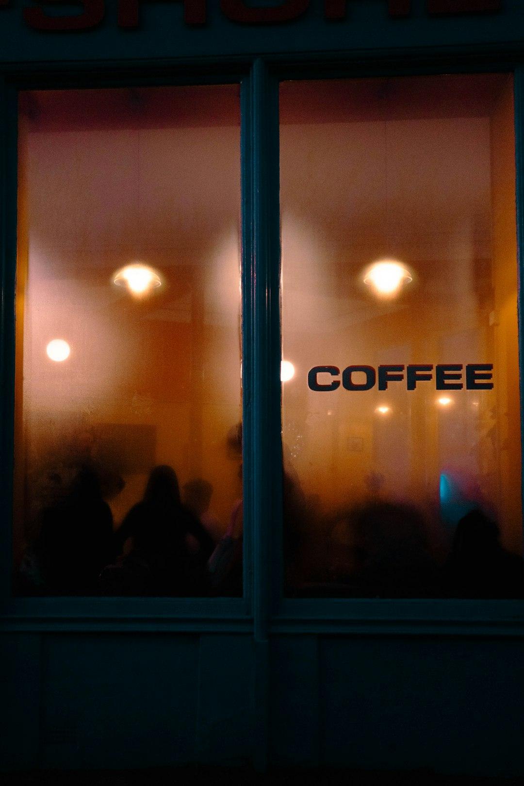 Kawiarnia nocą, ciepłe światło, sylwetki ludzi, napis 'COFFEE' na oknie.