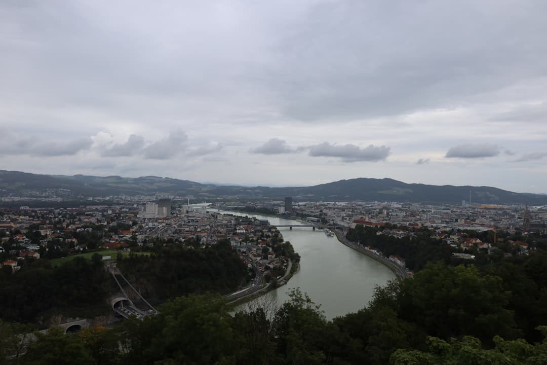 Panoramiczny widok miasta z rzeką, mostami i wzgórzami pod chmurami.