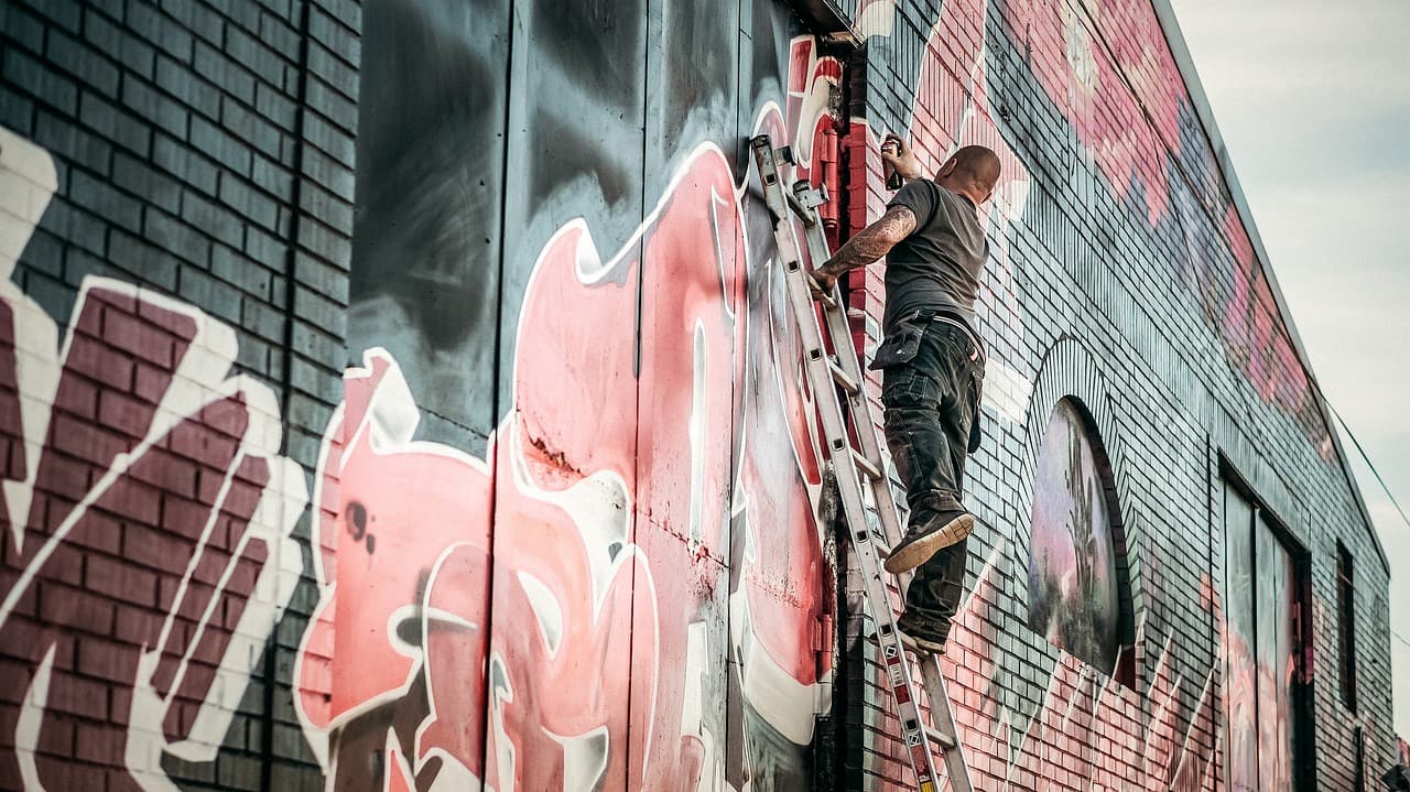 Mężczyzna malujący kolorowe graffiti na ceglanej ścianie z drabiny.