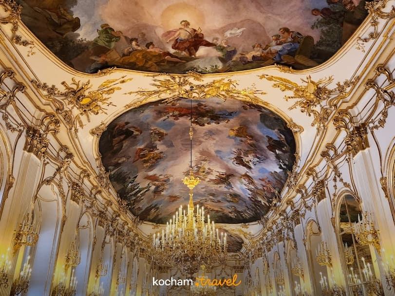 Opulentne wnętrze pałacu, złote sztukaterie, freski sufitowe, kryształowe żyrandole.