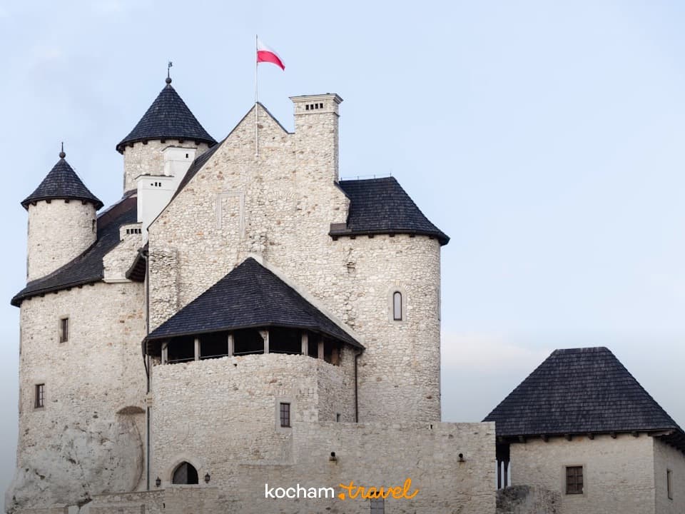 Zamek w Pieskowej Skale, białe ściany, czarne dachy, polska flaga.