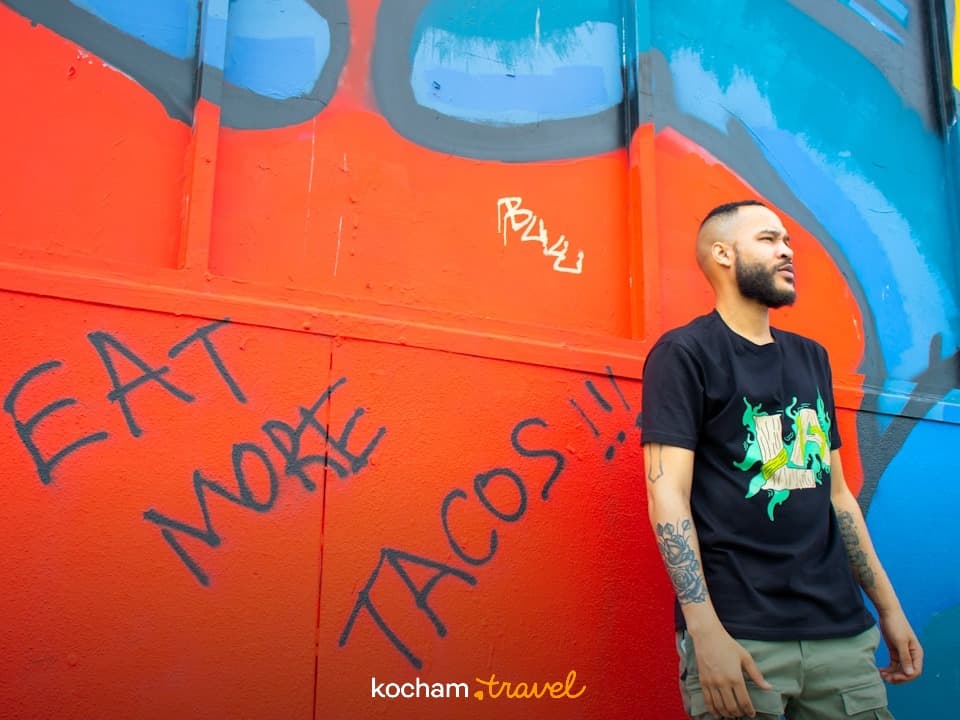 Mężczyzna przed kolorową ścianą z graffiti "EAT MORE TACOS".