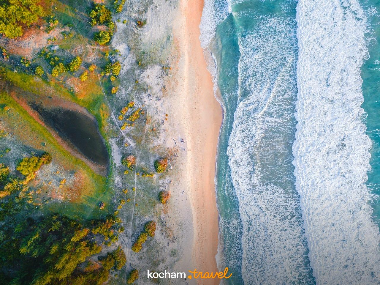 Widok z lotu ptaka: piaszczysta plaża, turkusowy ocean, fale, roślinność przybrzeżna, laguna.