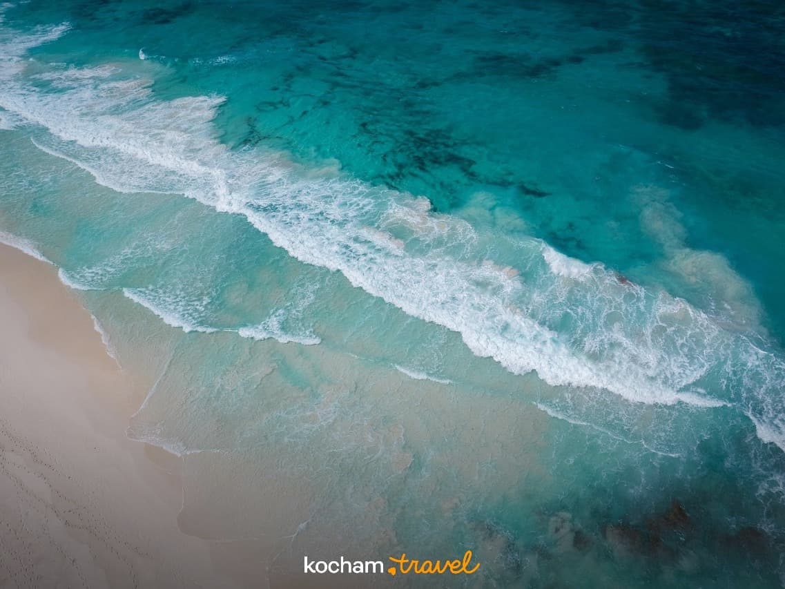 Turkusowe fale oceanu rozbijające się o piaszczystą plażę z lotu ptaka.