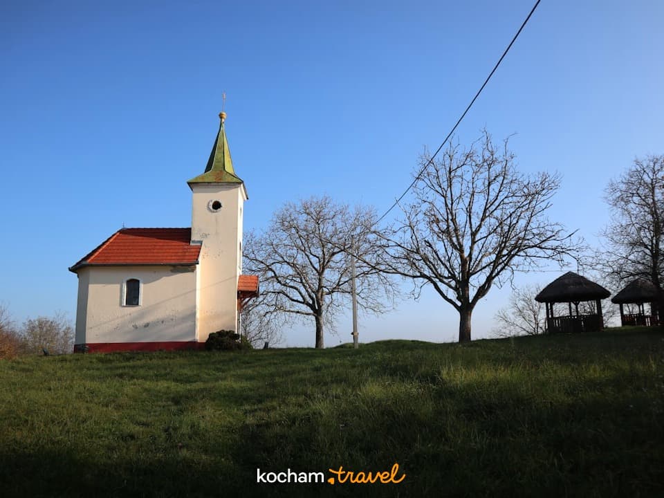Mały biały kościół, zielone wzgórze, suche drzewa, błękitne niebo, altanki.