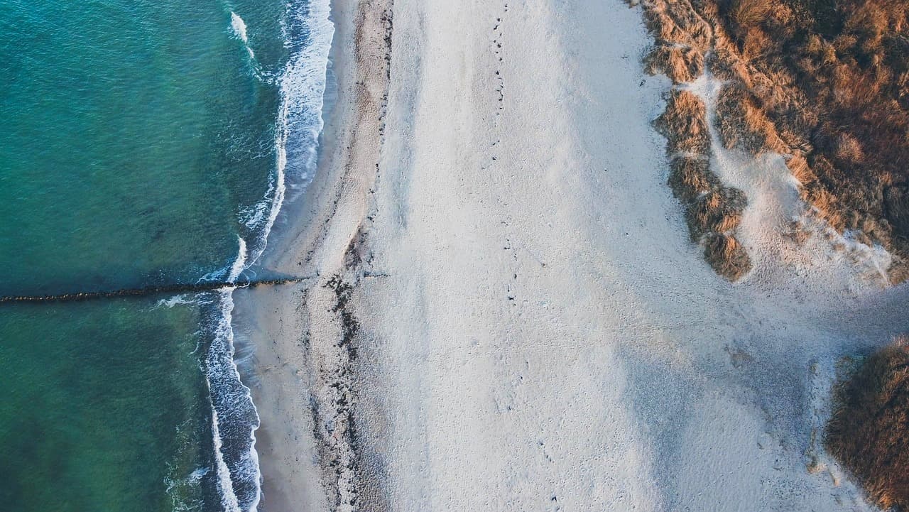 Widok z lotu ptaka: turkusowy ocean, fale, piaszczysta plaża, wydmy, falochron.