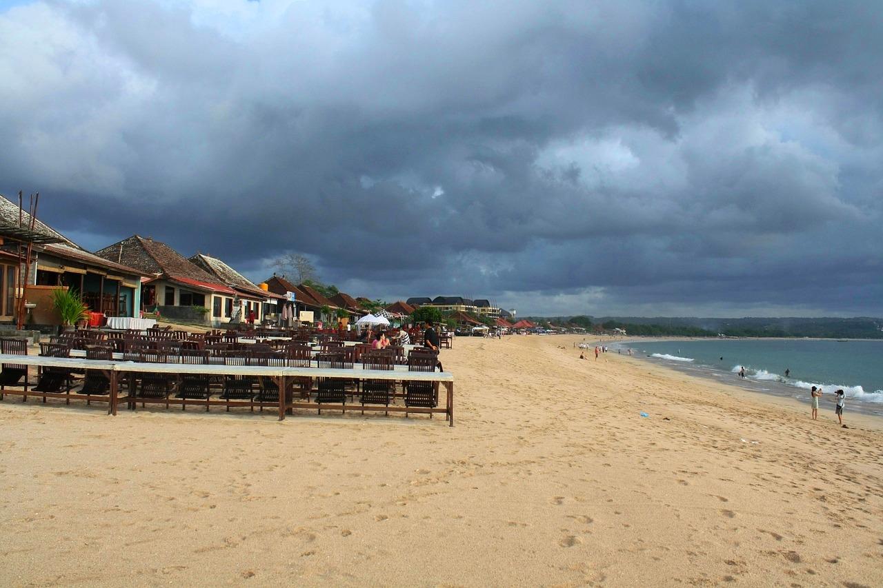 Piaszczysta plaża z restauracjami, turkusową wodą i ciemnymi chmurami.