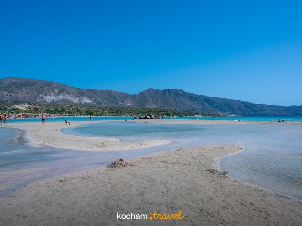 Plaża z turkusową wodą, różowym piaskiem i górami w tle.