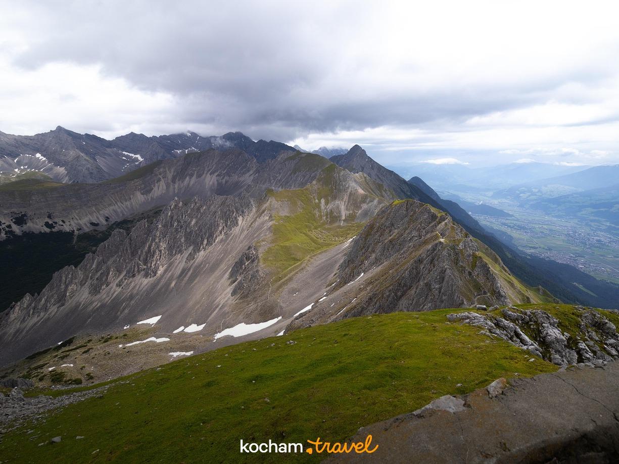 Panoramiczny alpejski krajobraz, skaliste szczyty, zielone zbocza, chmury.