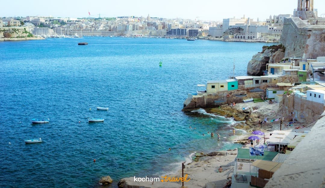 Malta, Valletta, skaliste wybrzeże, turkusowa zatoka, miasto, łodzie i kąpiący się ludzie.