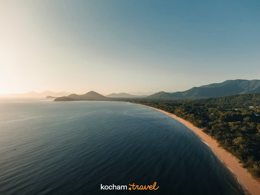Panoramiczny widok na długą piaszczystą plażę, ocean, las, góry, zachód słońca.