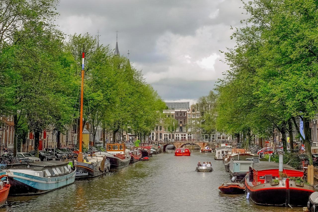 Kanał w Amsterdamie z łodziami, zielonymi drzewami i zabytkowymi budynkami.