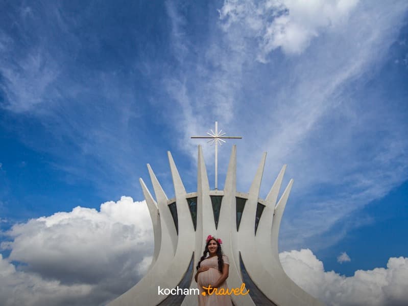 Kobieta w ciąży pozuje przed Katedrą w Brasílii, modernistyczna architektura, błękitne niebo.