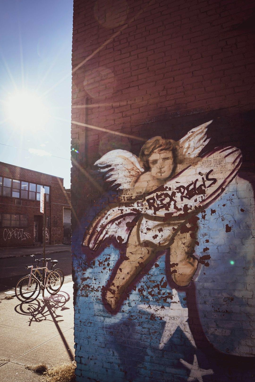 Graffiti anioła Rex Regis na ceglanej ścianie, rowery na słonecznej ulicy.
