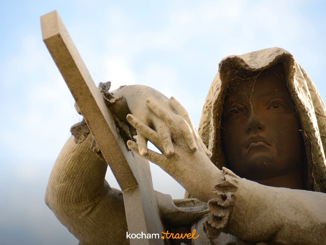 Zwietrzała kamienna statua postaci w kapturze z krzyżem.