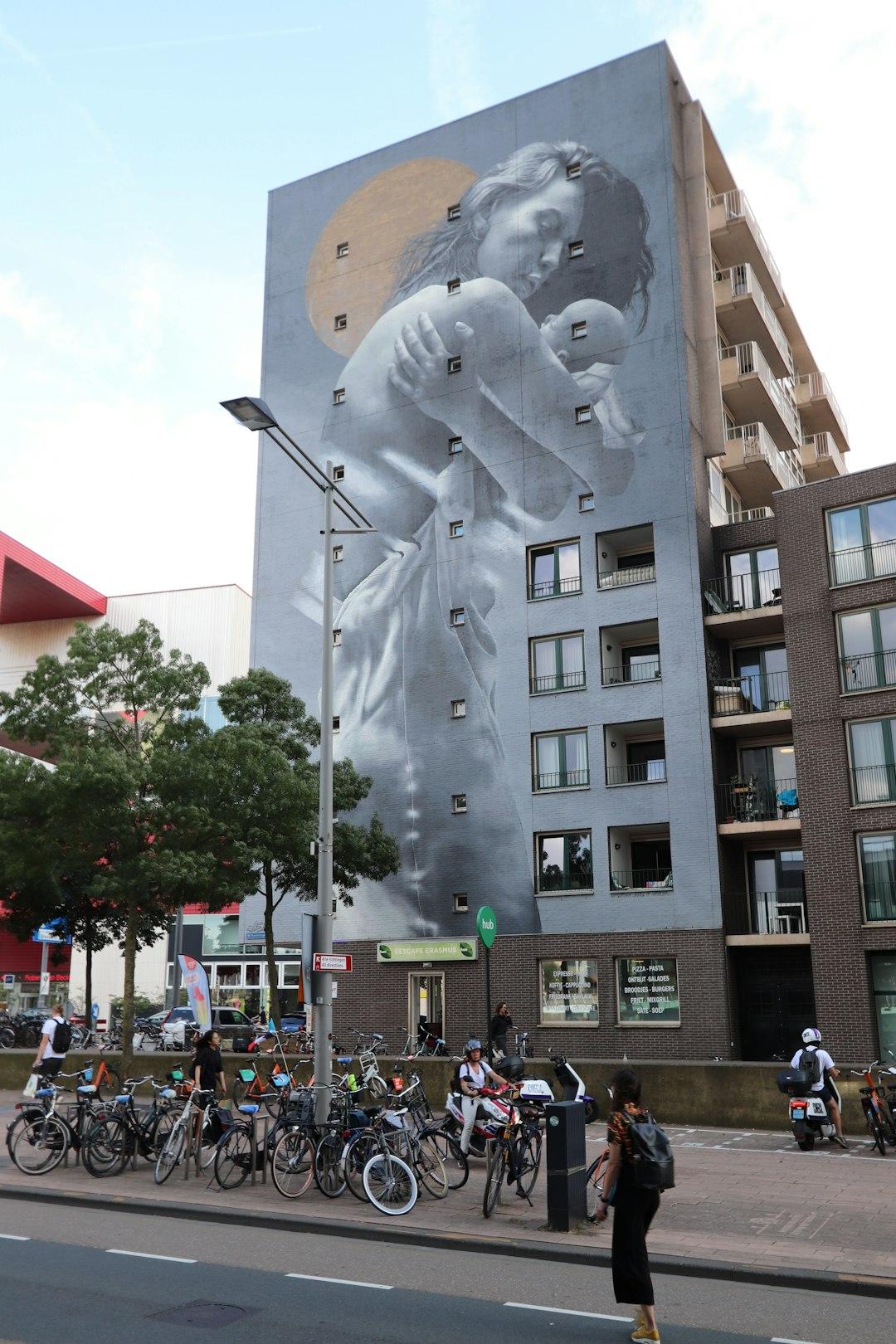 Duży mural kobiety z dzieckiem na ścianie budynku, ruchliwa ulica z rowerami.