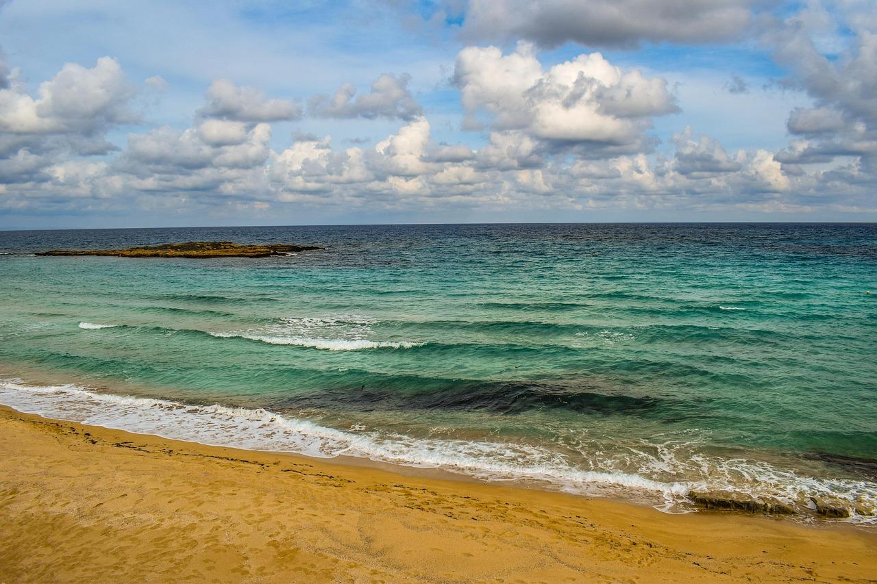 Plaża, turkusowe morze, fale, skalista wyspa, chmurne niebo.