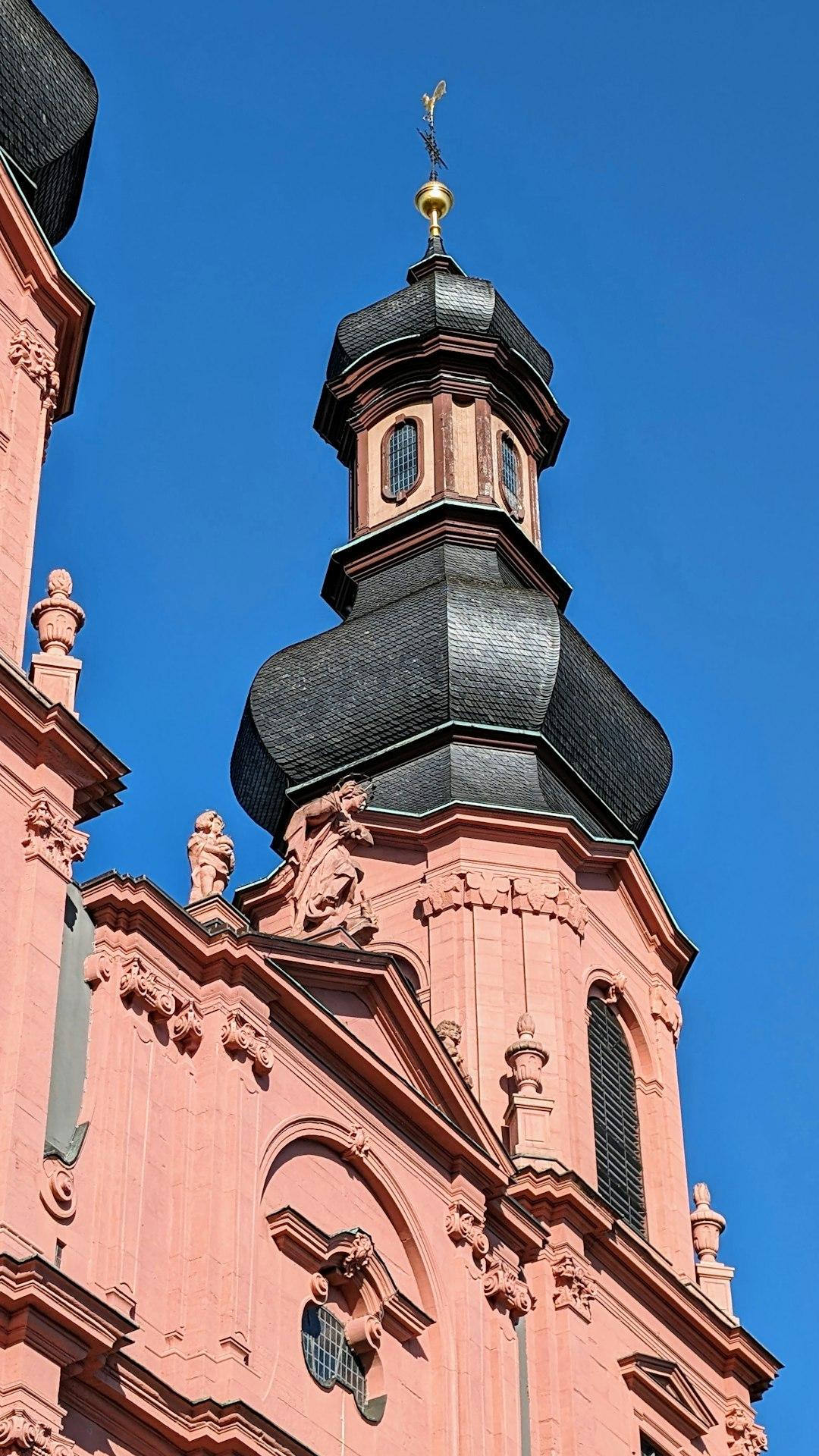 Różowa barokowa wieża kościelna z ozdobnym dachem i złotą iglicą.
