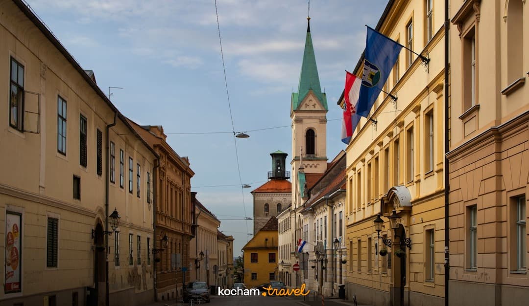 Zabytkowa ulica, stare budynki, wieża kościelna, zielona iglica, chorwackie flagi, słoneczny dzień.