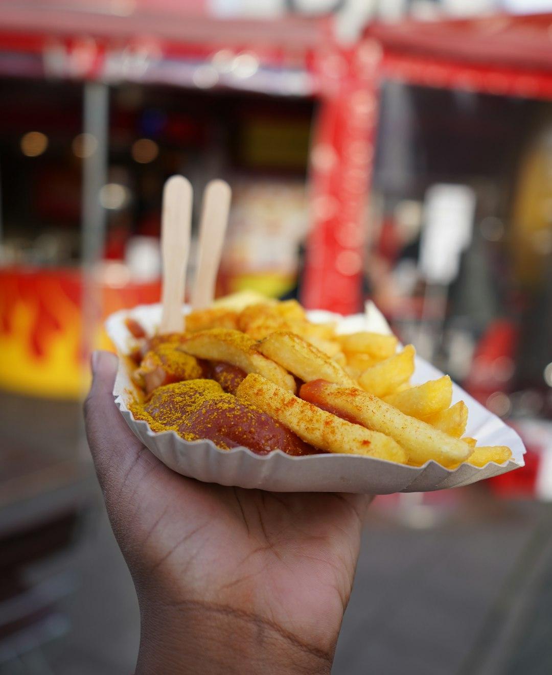 Currywurst z frytkami i sosem curry trzymane w ręku na ulicy.