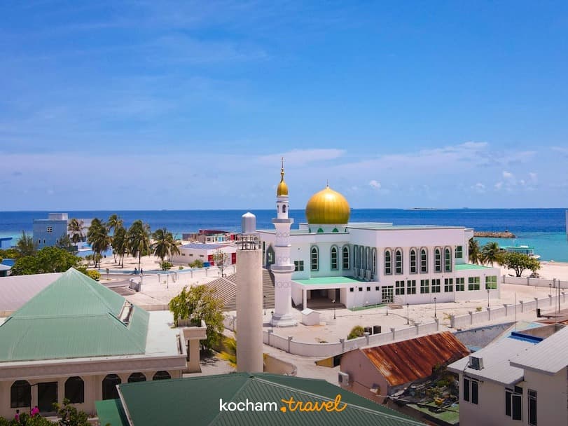 Biały meczet ze złotą kopułą, minaretem, lazurowym oceanem i palmami.