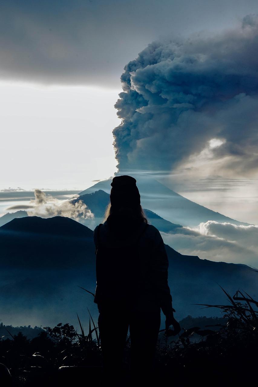 Sylwetka osoby obserwującej erupcję wulkanu z dymem nad górami.