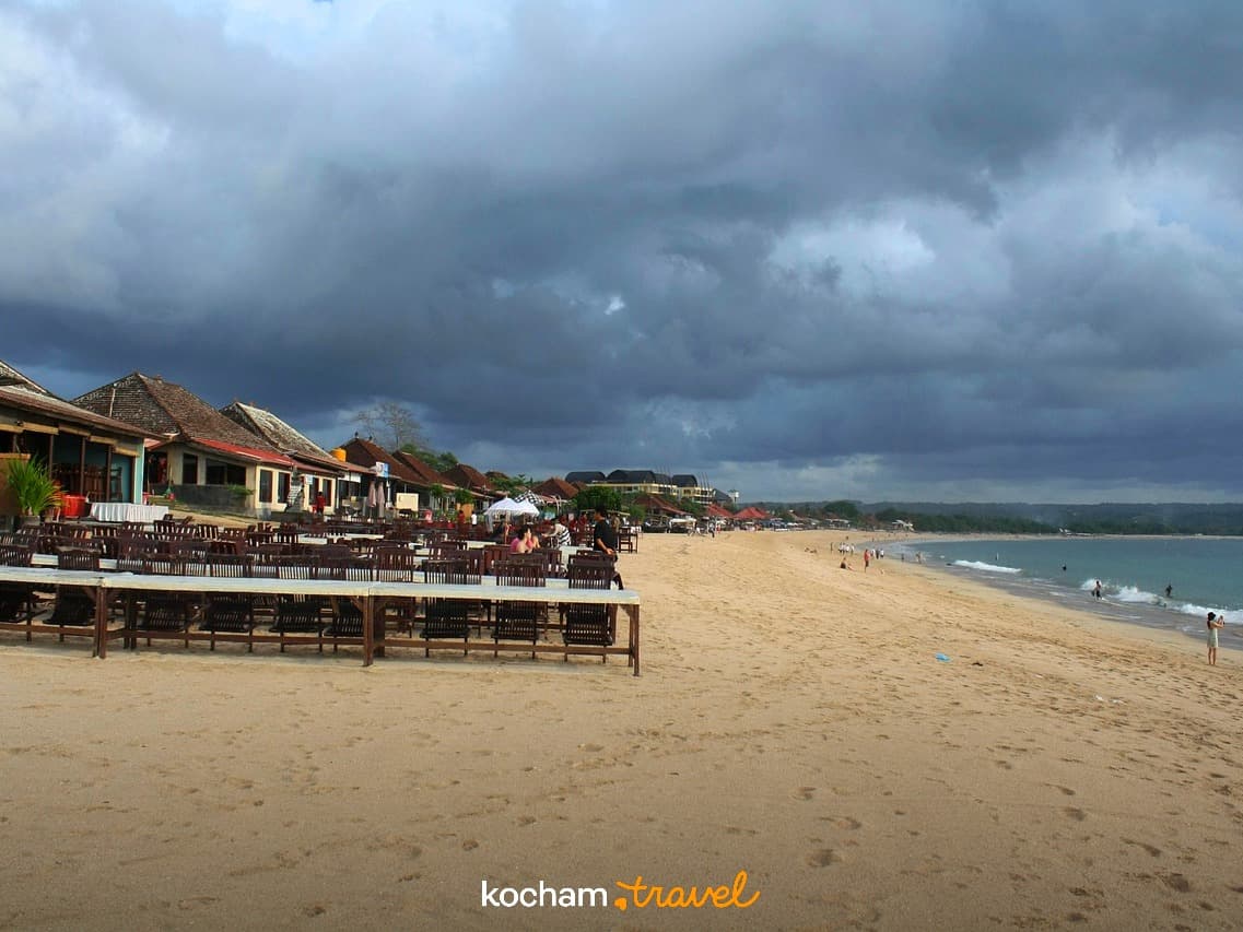 Szeroka piaszczysta plaża z restauracjami i turkusowym morzem pod ciemnymi chmurami.