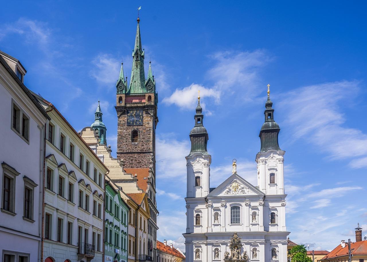 Kamienna wieża zegarowa, biały kościół barokowy, kolorowe kamienice, błękitne niebo.