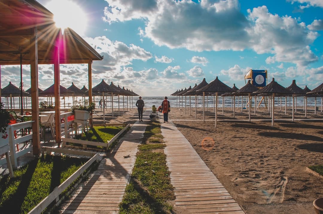 Ludzie spacerujący drewnianą ścieżką na słonecznej plaży z parasolami.