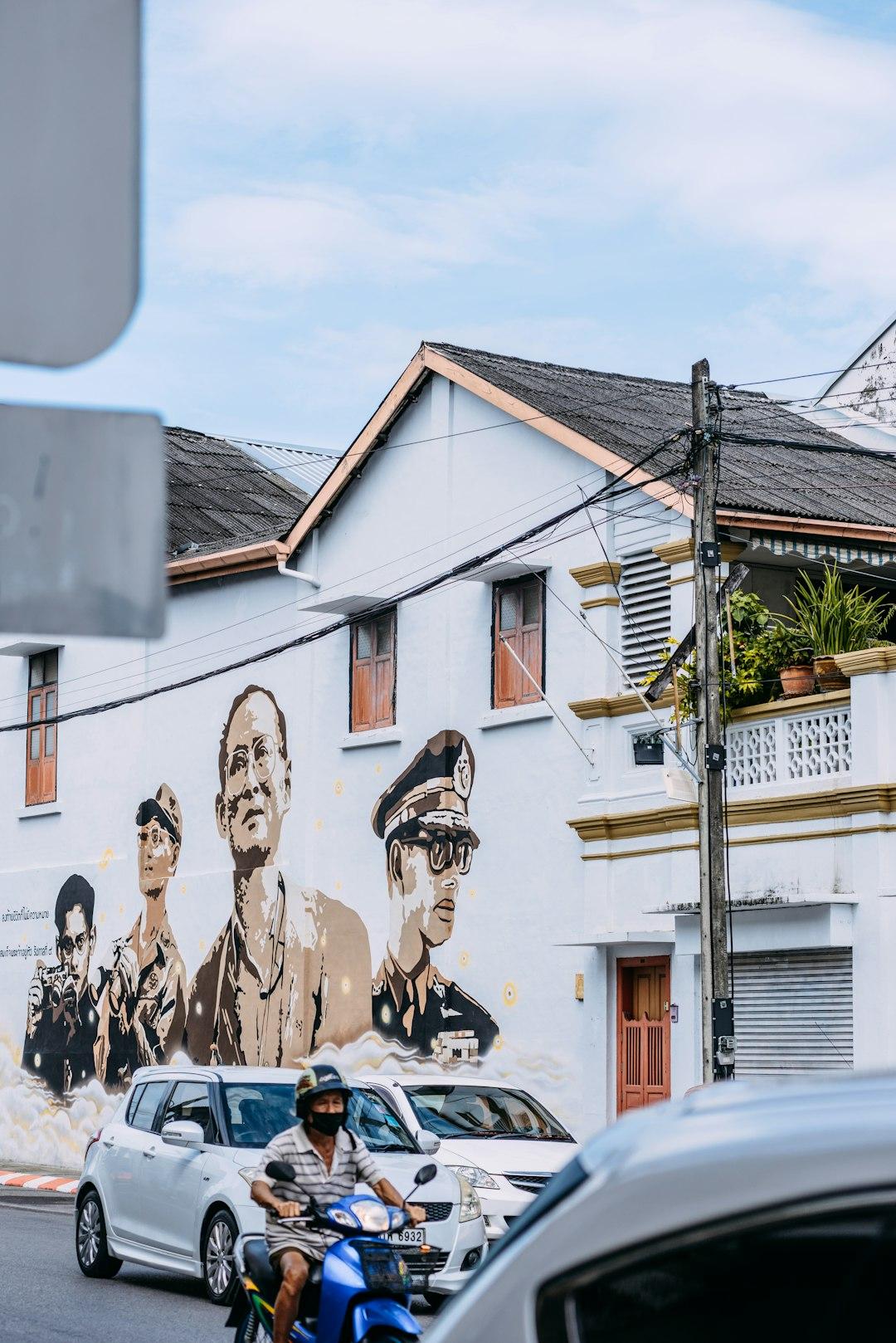 Mural z portretami na białym budynku, ulica, skuter, samochody.