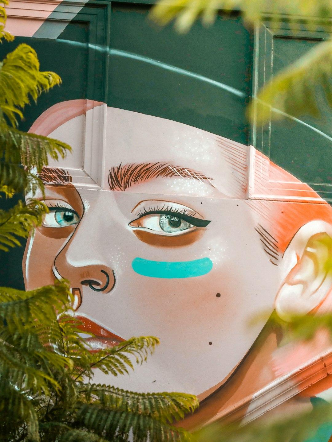 Sztuka uliczna: mural twarzy z turkusowym paskiem, otoczony zielenią.