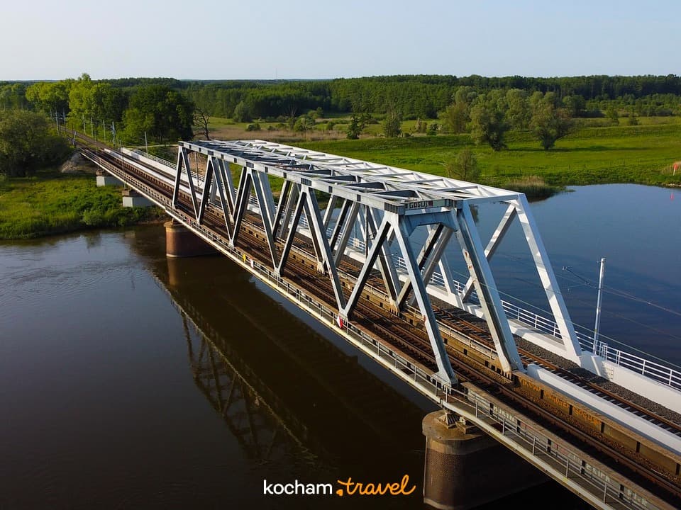 Metalowy most kolejowy nad rzeką, zielone pola i lasy.