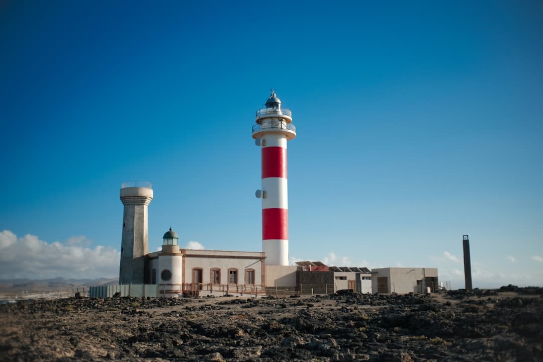 Czerwono-biała latarnia morska na skalistym wybrzeżu pod błękitnym niebem.