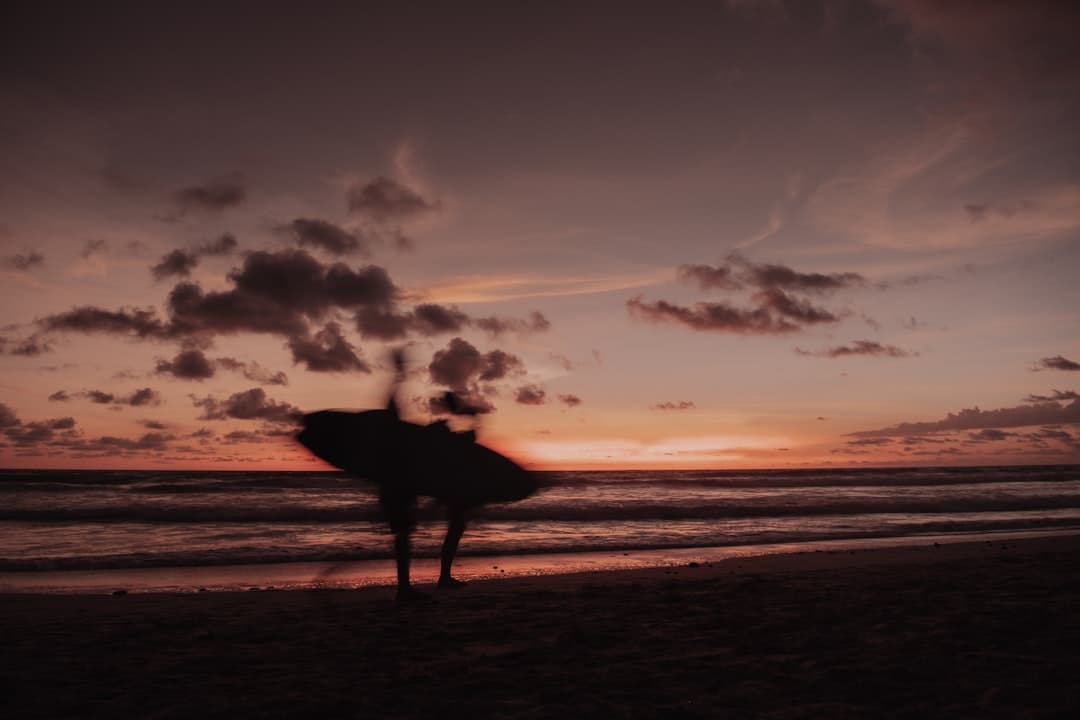 Rozmazana sylwetka osoby z deską surfingową na plaży o zachodzie słońca.