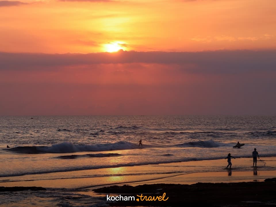 Ciepły zachód słońca nad oceanem, sylwetki ludzi na plaży i surferzy.