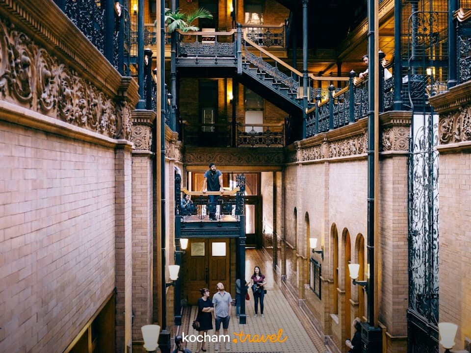 Wnętrze zabytkowego budynku: atrium, żeliwne balustrady, ceglane ściany, ludzie.