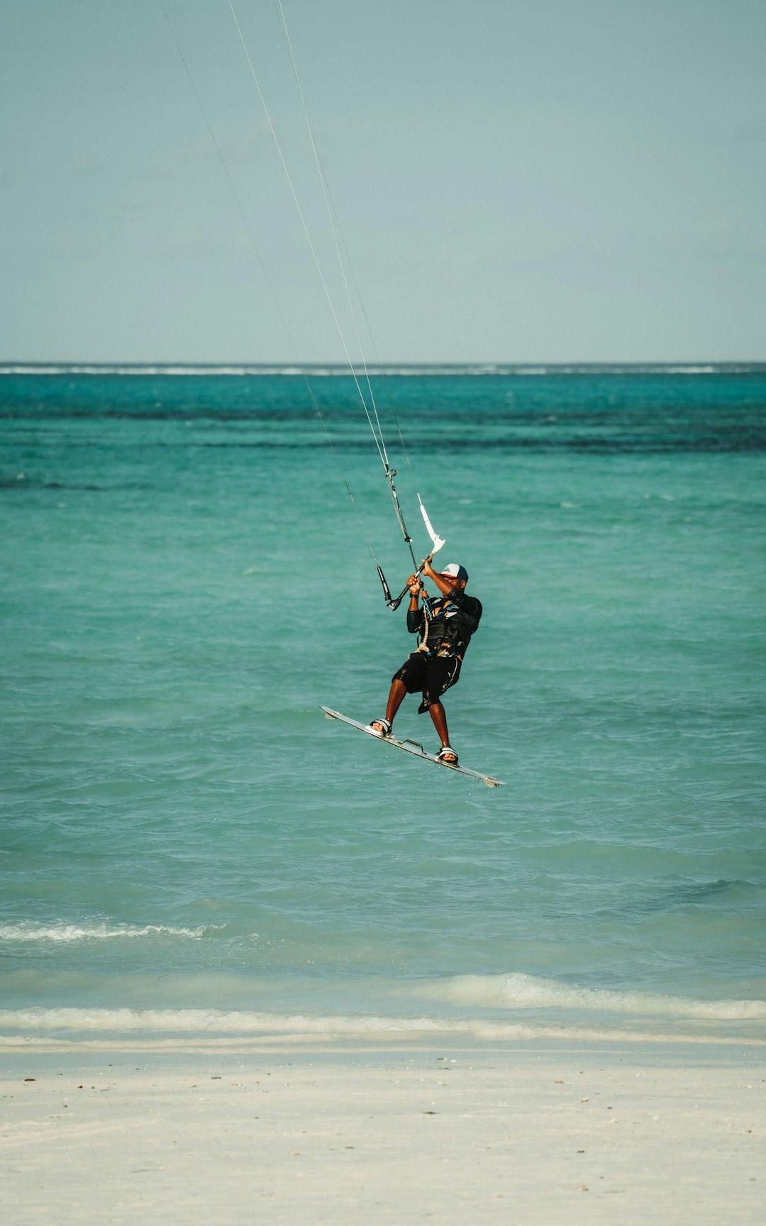 Kitesurfer wyskakujący z turkusowej wody na tle piaszczystej plaży.