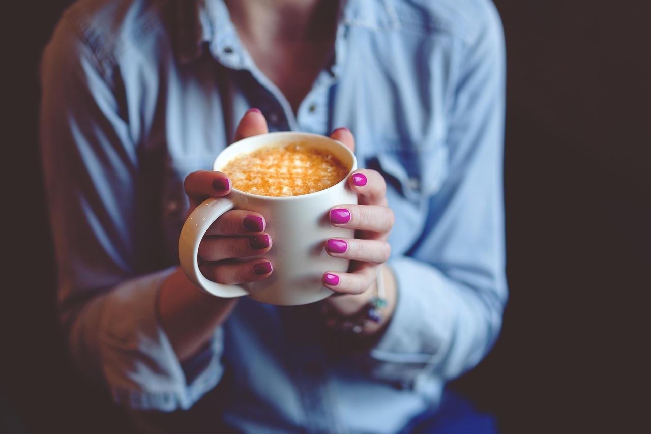 Kobieta z różowymi paznokciami trzymająca biały kubek kawy latte.