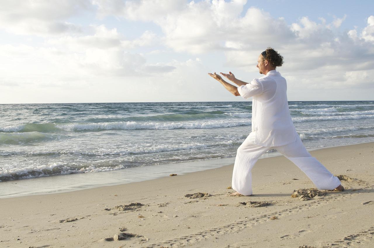 Mężczyzna w białym ubraniu ćwiczący Tai Chi na piaszczystej plaży.