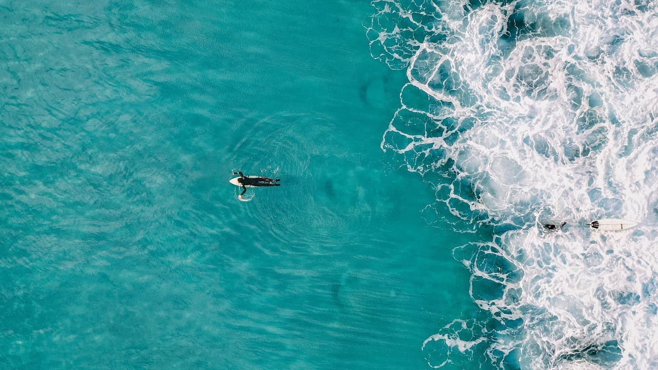 Widok z góry: surferzy, turkusowy ocean, białe fale, słoneczny dzień.