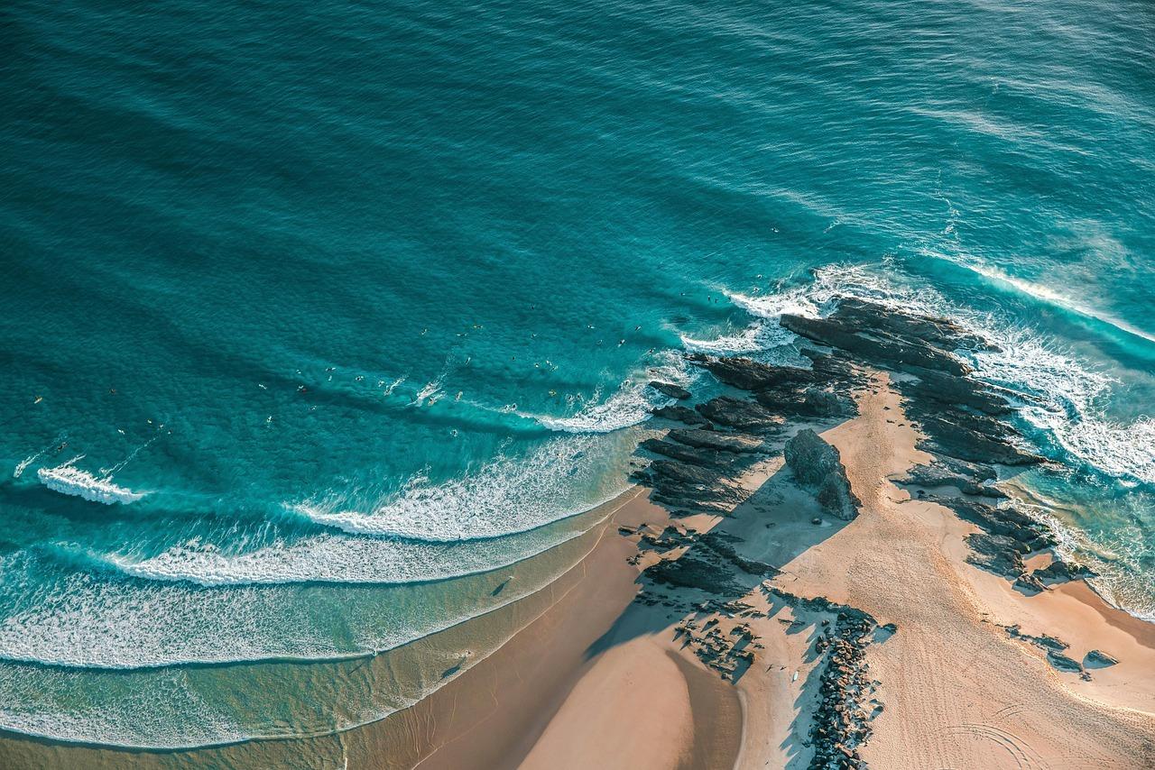 Słoneczny widok z lotu ptaka: plaża, turkusowy ocean, surferzy, skały.