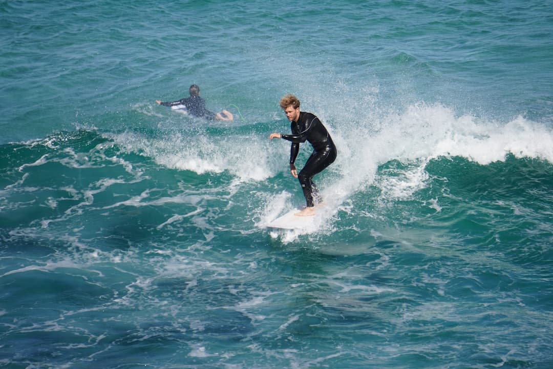 Dwóch surferów w piankach, jeden łapie falę, drugi czeka.