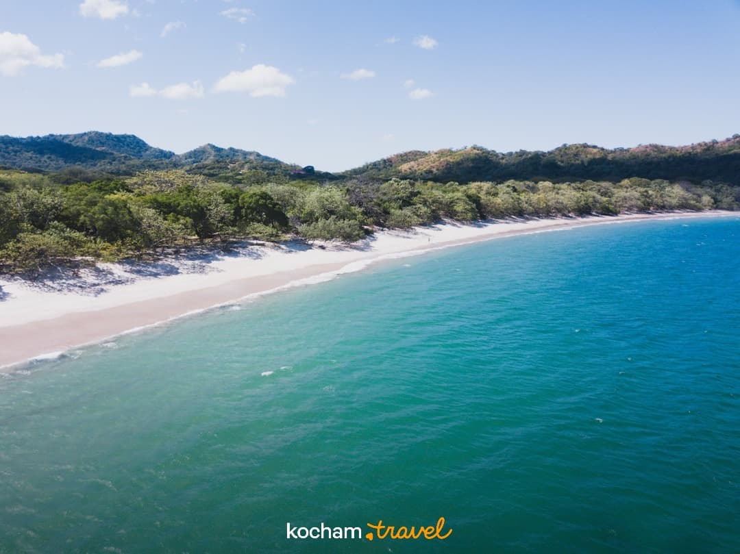 Piaszczysta plaża, turkusowa woda oceanu, tropikalny las, wzgórza.