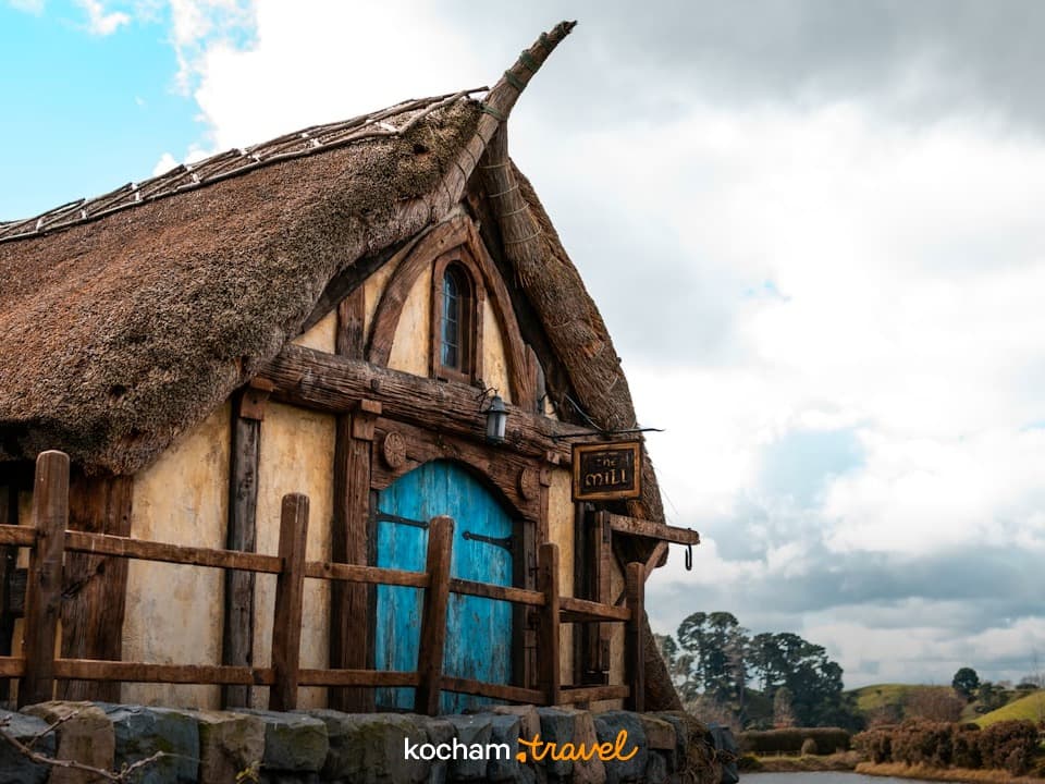 Młyn z niebieskimi drzwiami i dachem ze strzechy w krajobrazie fantasy.
