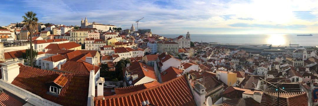 Panorama Lizbony, dachy z terakoty, zabytkowe budynki, odbicie słońca na wodzie.